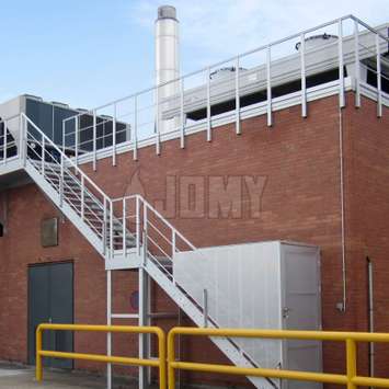 Escaleras de acceso a maquinaria con puerta de seguridad y barandillas para la protección colectiva contra caídas en el techo de un edificio de una fábrica.