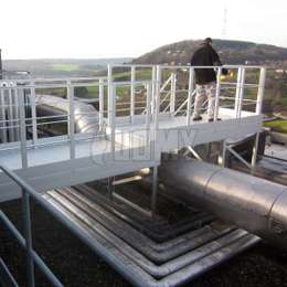 Pasarela de aluminio en techo plano usada para caminar sobre una tubería hasta llegar a una escalera metálica. 