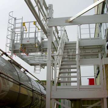 Platform accessed by stairs and equipped with cage ladders for accessing tanker truck manholes.